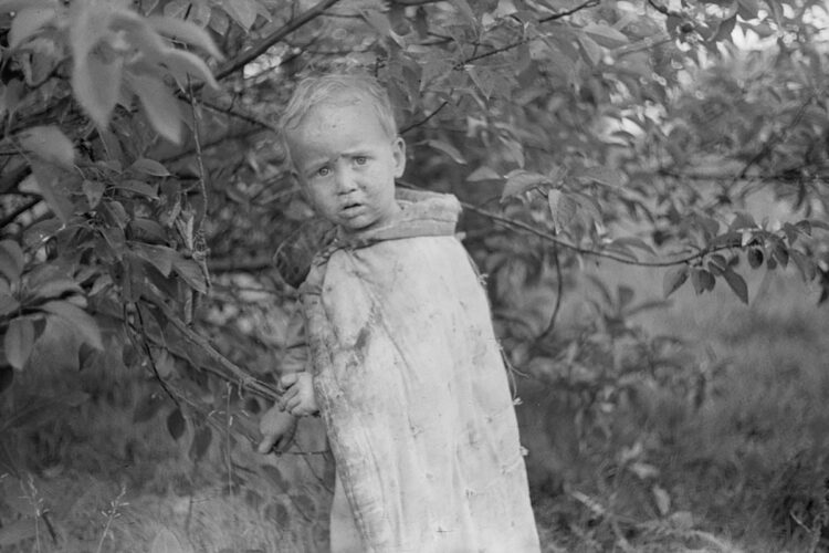 Great Depression era black & white photo of a small child has a blanket pinned about him in place of a jacket as he pokes among a large bush, accompanying his family who are picking berries in Berrien Springs, Michigan